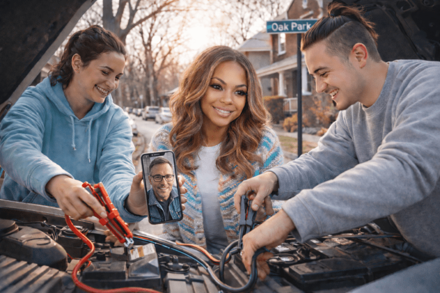 Dr. Tracey Bond stands between two late-twenties neighbors in hoodies as they help jump-start her Range Rover Evoque on an Oak Park, Illinois street. One neighbor holds jumper cables while the other connects them to the battery under the open hood. On a cell phone screen, a middle-aged man with glasses appears via video call offering guidance. The group smiles warmly, capturing a moment of community, collaboration, and neighborly kindness.